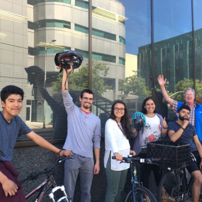 A group of six people with bicycles stands in front of a glass building. One person holds a helmet in the air, and another waves at the camera. They are all smiling and appear to be enjoying a sunny day. Trees and reflections are visible in the building's windows.