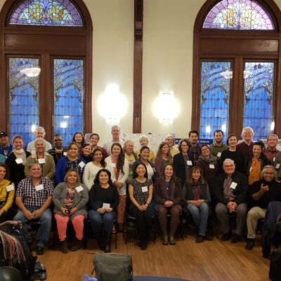 A diverse group of people gathered for a group photo in a large, warmly lit room with stained glass windows. Some individuals are seated while others stand behind them, all smiling at the camera. Tables with papers and name tags are visible in the foreground.