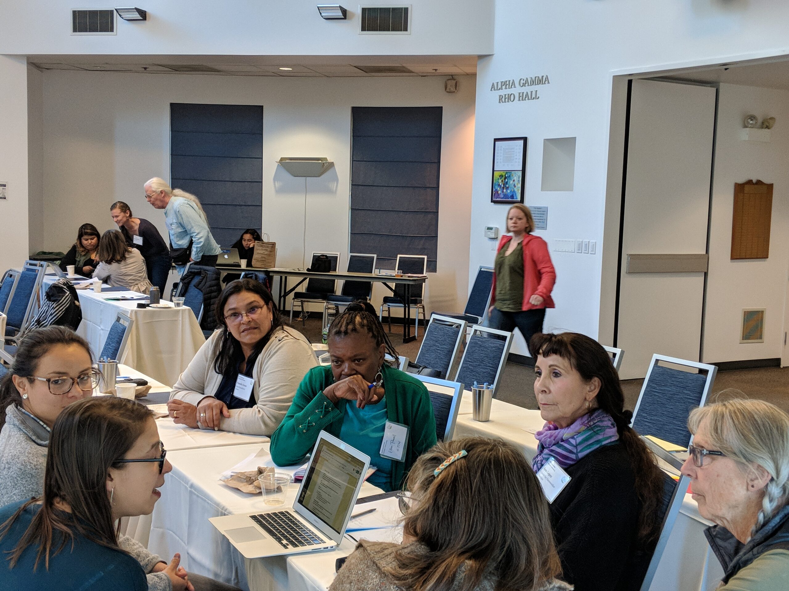 Un grupo de personas están sentadas a una mesa, enfrascadas en una discusión en una sala de conferencias llamada "Alpha Gamma Rho Hall". Algunos toman notas en sus ordenadores portátiles y otros escuchan atentamente. Al fondo se ve a otros asistentes, uno de pie y otro caminando.