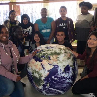 A diverse group of people are gathered around a large globe of Earth. They are smiling and appear to be in a classroom or educational environment with natural decorations in the background. Some are standing while others kneel or sit around the globe.