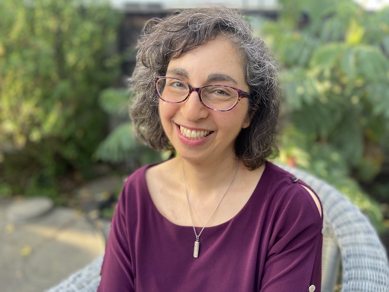 Fotografía de una mujer sonriente con camisa morada y gafas delante de un fondo verde.
