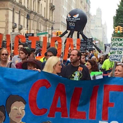 A large group of people are marching in a city street, holding banners and signs. The prominent banners include messages like "California," "Fossil Free," and "Divest!" Some people are wearing colorful clothing and hats, and buildings line the street in the background.