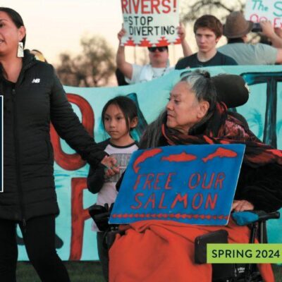 A person speaks into a microphone at an outdoor event with signs advocating for river conservation and salmon freedom, attended by people of various ages.