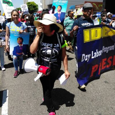 A large group of protestors march down a city street carrying various signs and banners. A woman wearing a wide-brim hat, sunglasses, and holding a megaphone leads the group. The banners behind her promote workers' rights and day labor programs.