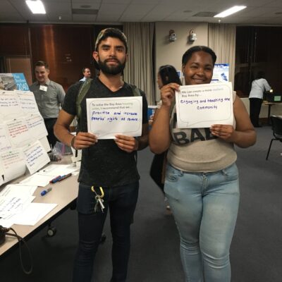 Two individuals stand side by side in a room, each holding a small whiteboard with messages written on them. The person on the left's board reads, "Prevent and increase density right at market," and the person on the right's board reads, "Engaging and teaching the community.