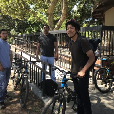 Three people are standing near bicycles. One person on the right, wearing a bike helmet, is holding a bike. The two others are standing, engaged in conversation. There is a railing and some greenery in the background. It appears to be a sunny day.