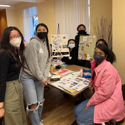 A group of diverse people, wearing face masks, pose together in a room. Some are seated at a table covered with creative materials like markers and paper, while others stand behind it. They showcase their collaborative artwork, featuring various cutouts and designs.