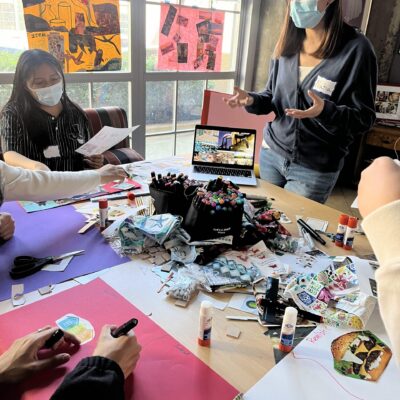 A group of people are gathered around a table filled with art supplies, including markers, glue sticks, and paper. Two individuals wear masks and engage in conversation. One person stands and gestures while others work on projects, surrounded by colorful posters and windows in the background.