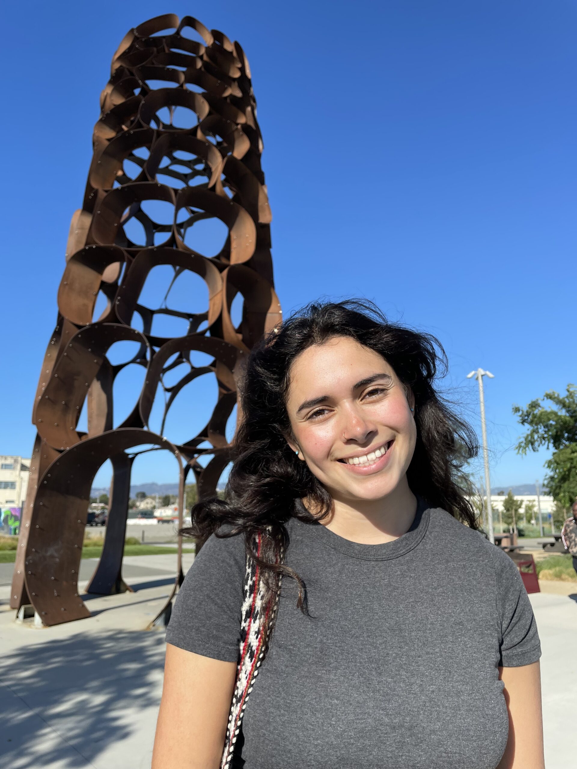 Mujer joven de pie delante de una obra de arte con camisa gris y pelo castaño.