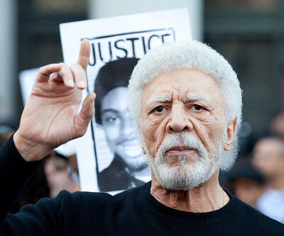 A man with white hair and beard speaks passionately, raising one finger. He stands in front of a blurred crowd. Behind him, a sign with a black-and-white photo and the word "JUSTICE" is visible. The atmosphere suggests a protest or rally.