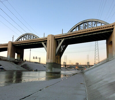 View of concrete-lined riverbed with two large, arched bridges spanning overhead. Power lines and towers are visible along the sides of the river. The sky is clear, with hints of dawn or dusk light.