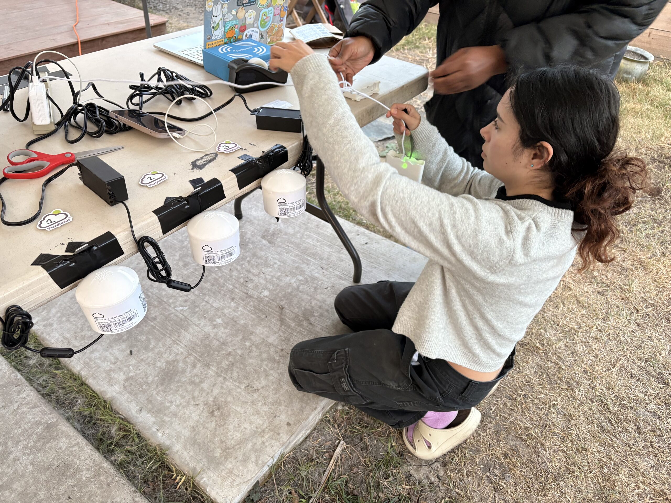 NVR_Staff_setting_up_AQ_monitors Young woman in white shirt sets up 3 air quality monitors on a white table.
