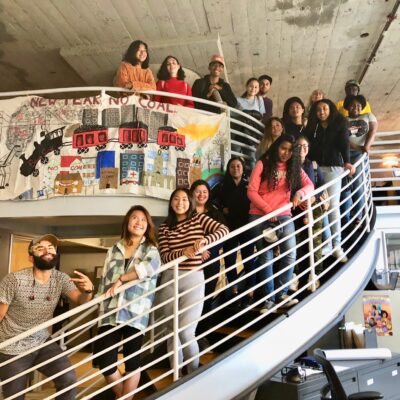 A group of 20 people standing and sitting on a spiral staircase indoors. They are smiling and posing for the photo. Behind them is a large banner with drawings and the text "Next year no coal." The setting appears to be an office or communal space.