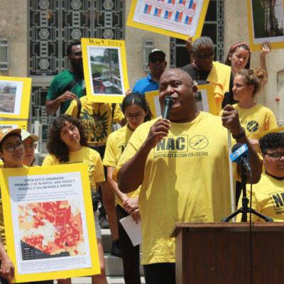 Residents in yellow t-shirts hold signs with images of flooding at a protest. A black man stands at a podium, speaking into a microphone in the foreground.