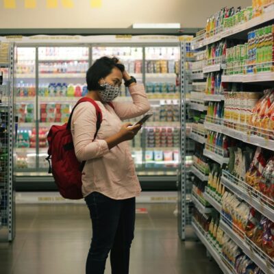 Una persona con una mascarilla y una mochila roja está de pie en el pasillo de un supermercado, con un teléfono móvil en la mano y rascándose la cabeza. Los estantes a ambos lados están repletos de aperitivos y productos diversos. El fondo muestra artículos refrigerados.