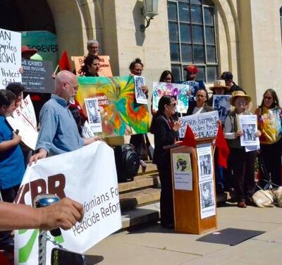 A group of people holding signs and banners are gathered outside a building for a protest. One person is speaking at a podium while others stand nearby, holding various posters advocating for pesticide reform.