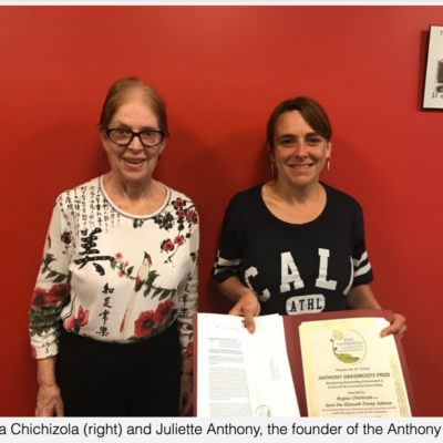 Two women standing against a red wall. The woman on the right holds a certificate and a folder, while the woman on the left smiles at the camera. Both are wearing glasses. A small framed picture with text hangs on the wall above their heads.