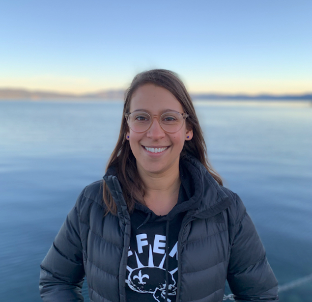 Woman with long brown hair and glasses stands in front of the ocean