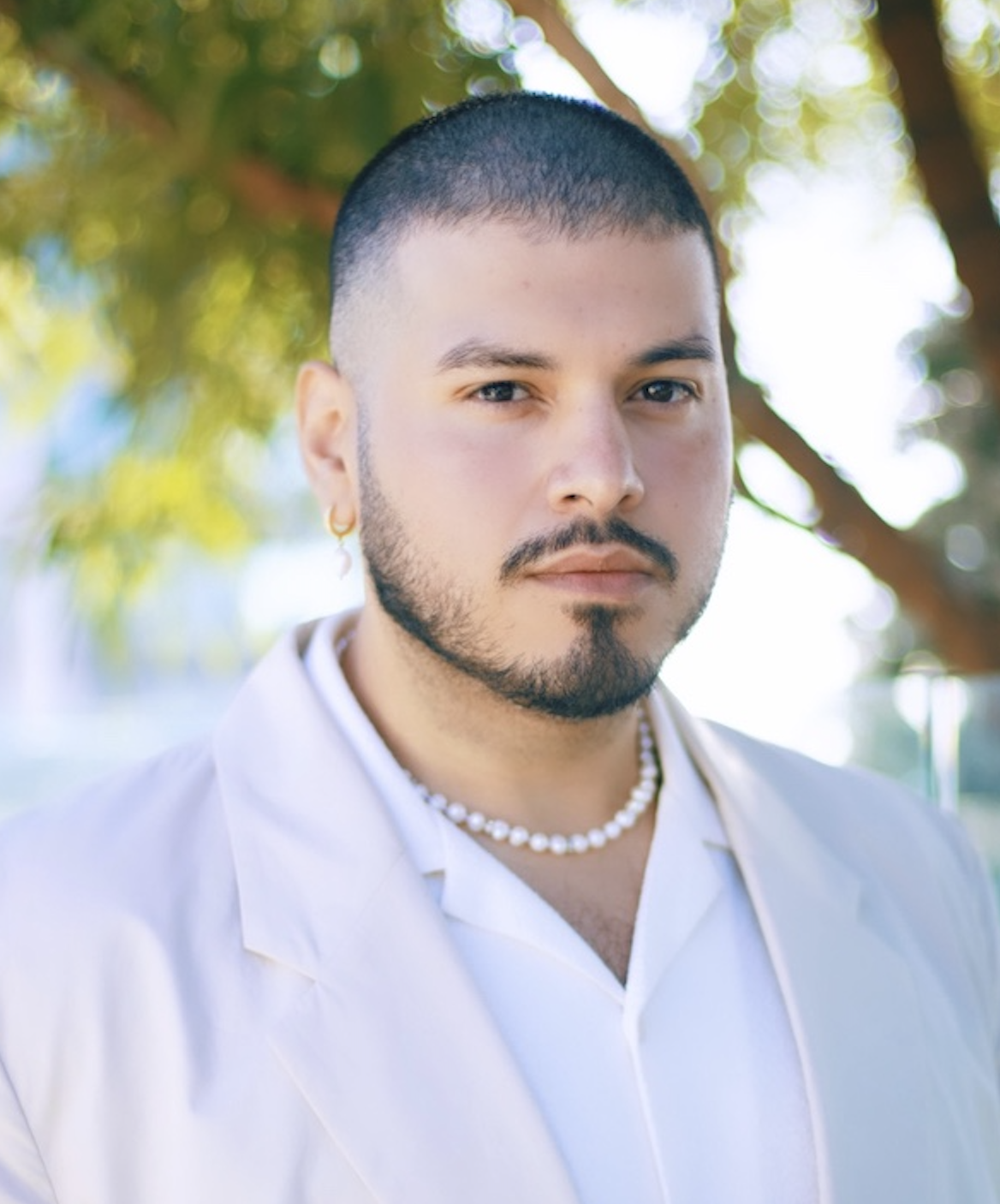 Fotografía de un hombre de pelo castaño con camisa y chaqueta blancas que lleva un collar de perlas.