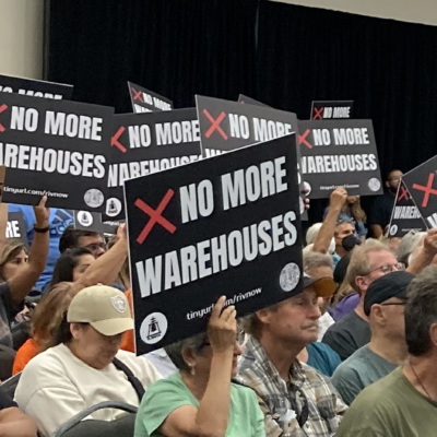 A crowd of people hold black signs that say "no more warehouses"
