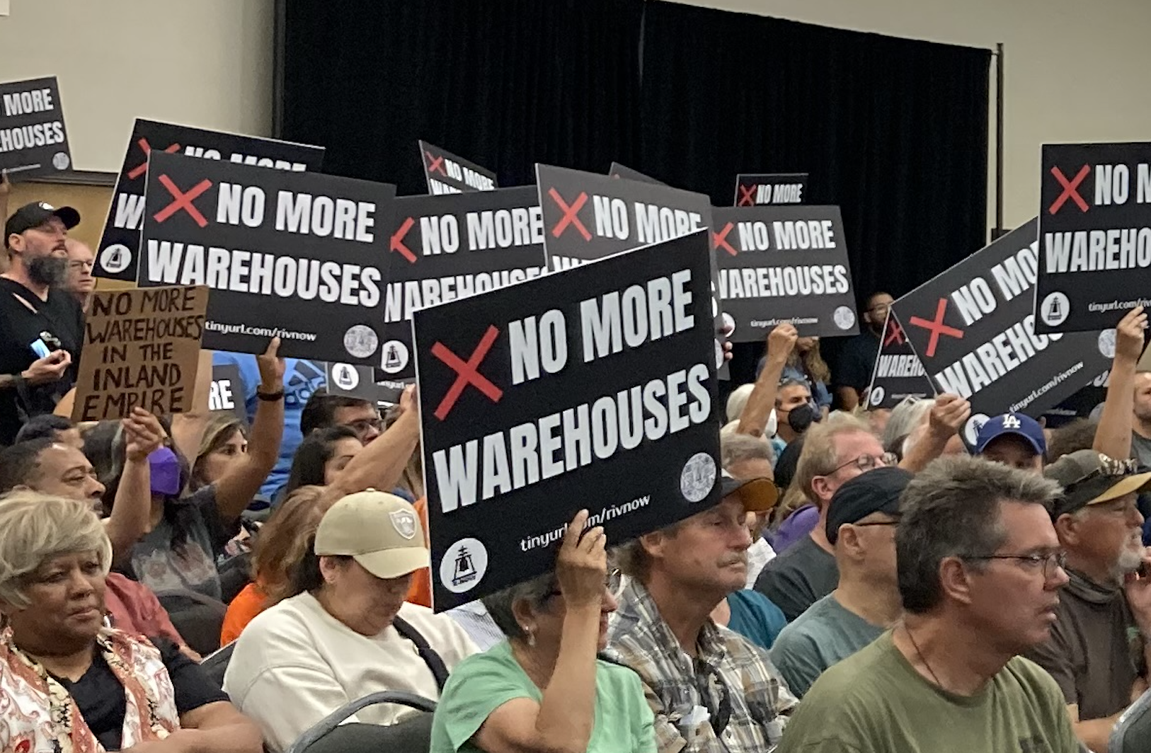 A crowd of people hold black signs that say "no more warehouses"