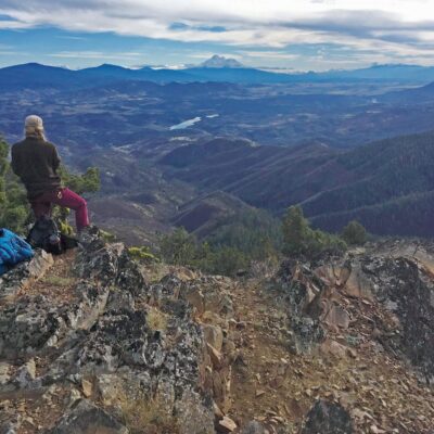 A person wearing a hat and jacket sits atop a rocky mountain peak, overlooking a vast landscape of rolling hills, valleys, and distant snowy peaks under a cloudy sky. A blue backpack rests beside them amidst the natural scenic view.