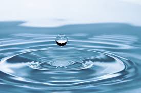 A single water droplet is suspended in mid-air above a body of water, creating concentric ripples on the water surface below. The image captures the moment just before the droplet makes contact with the water again.