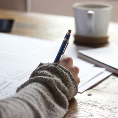 Una persona vestida con un jersey gris escribe con un bolígrafo azul sobre unas hojas de papel colocadas sobre una mesa de madera. En la mesa del fondo también hay una taza blanca y un cuaderno o una tableta.