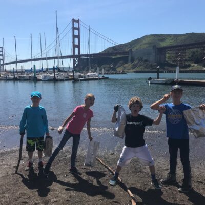 Four children stand on a beach holding bags and sticks, with the Golden Gate Bridge and docked sailboats in the background. The weather is sunny, and the children appear excited and enthusiastic, suggesting they might be participating in a cleanup activity.