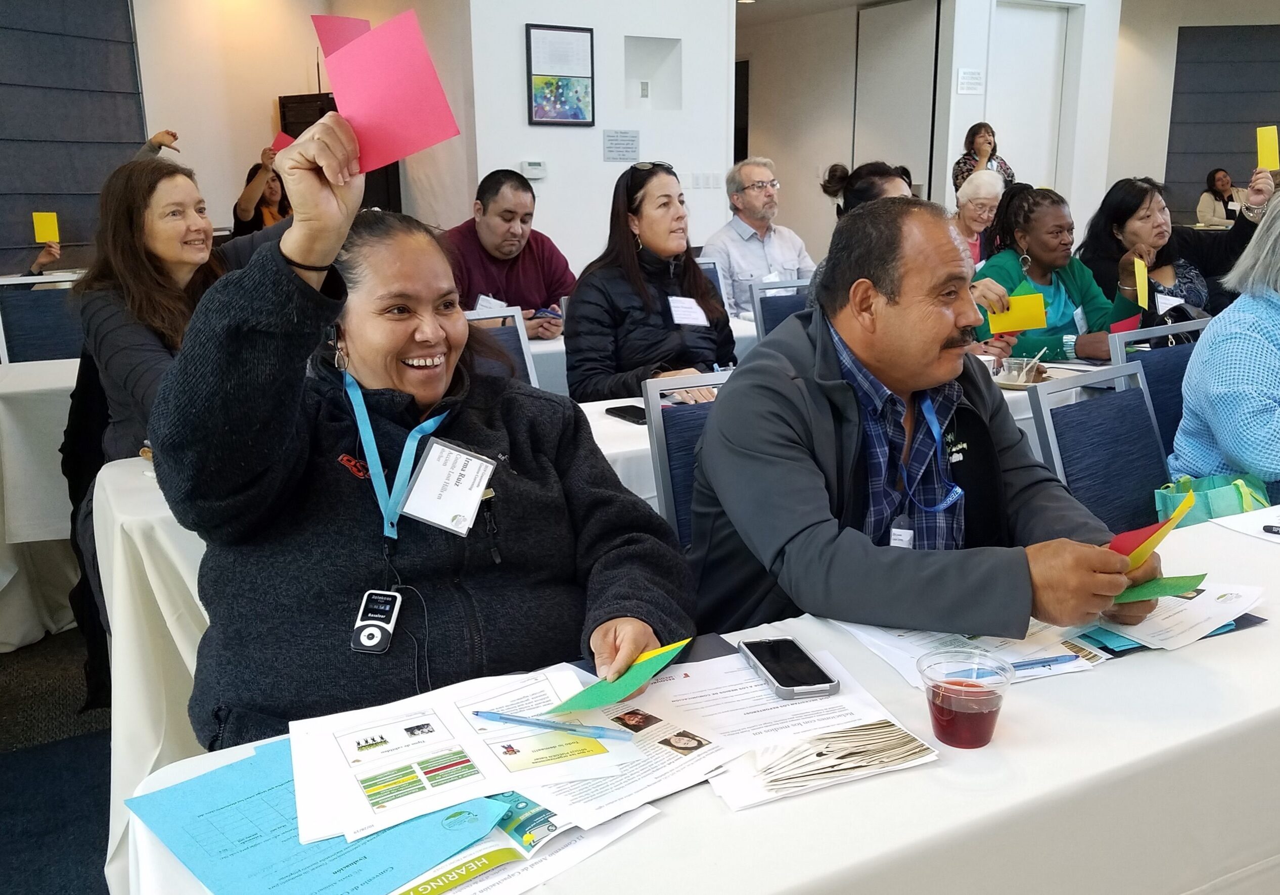 Un grupo heterogéneo de hombres y mujeres se sientan en mesas de conferencias y levantan tarjetas de colores. Una mujer sonriente a la izquierda sostiene una tarjeta rosa, mientras que un hombre a su lado levanta una tarjeta amarilla. Sobre las mesas se ven papeles, folletos y una bebida.