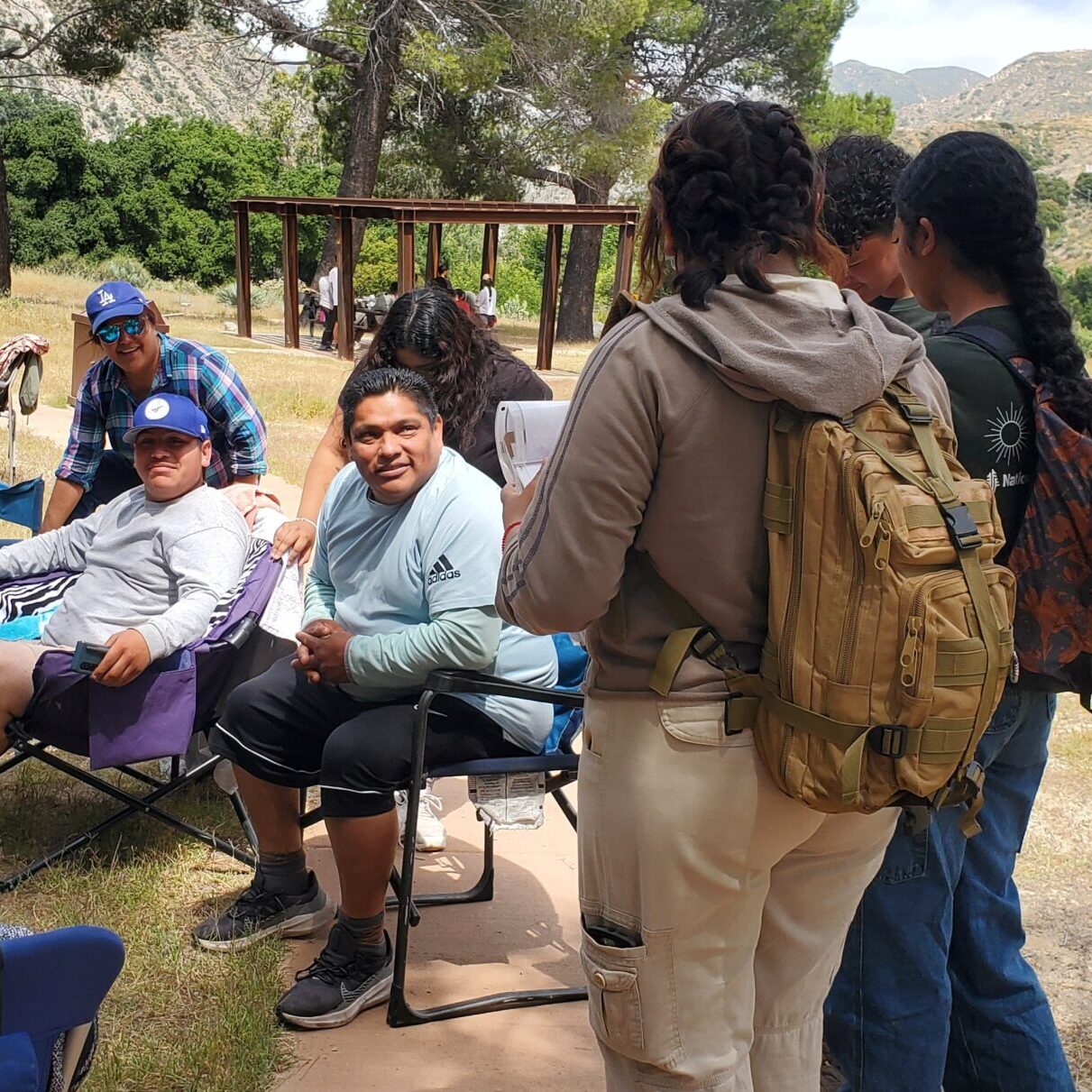 A group of people sitting and standing outdoors in a park area, with trees and mountains in the background.