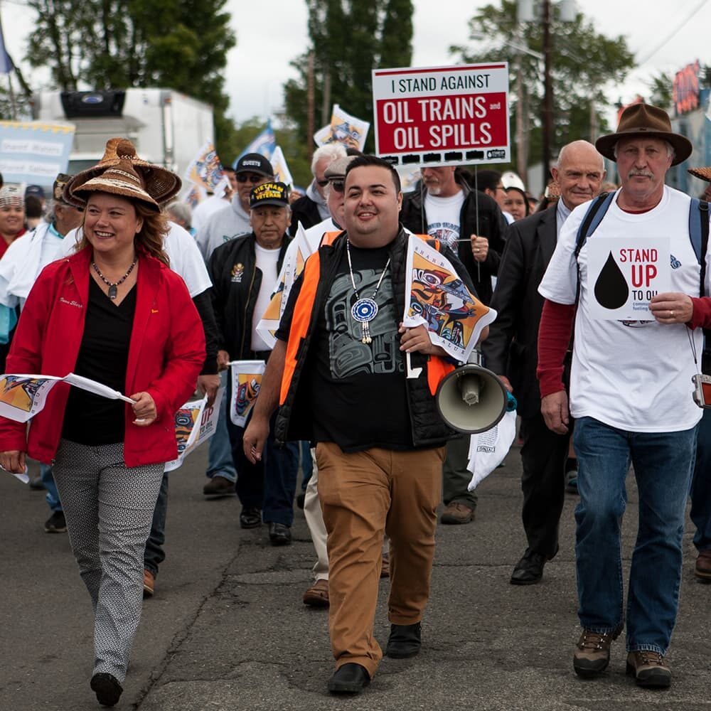 A diverse group of people participate in a protest march. Several hold signs, including one that reads "I STAND AGAINST OIL TRAINS AND OIL SPILLS." Some wear T-shirts with environmental messages, and one marcher carries a megaphone.