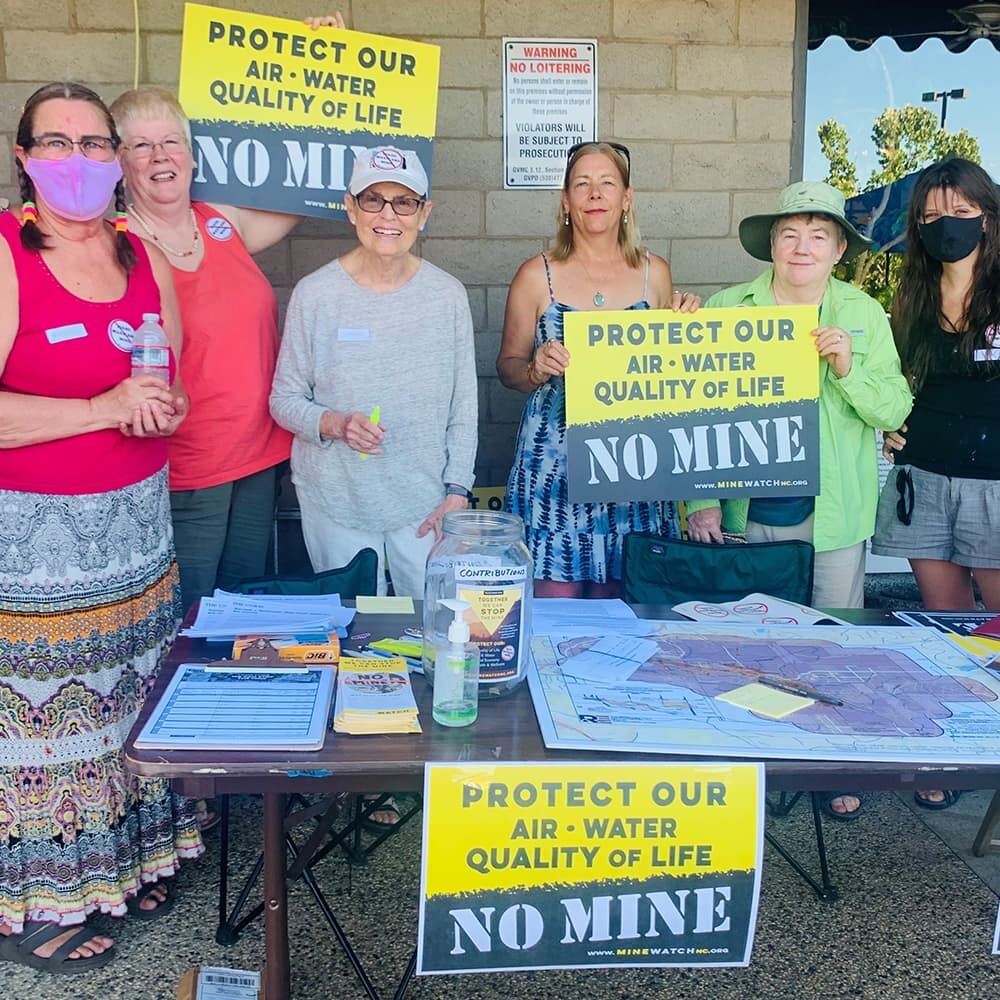 A group of people stands behind a table covered with flyers and a donation jar. They hold signs that say “No Mine” and “Protect Our Air, Water, Quality of Life.” They are participating in an environmental advocacy event.