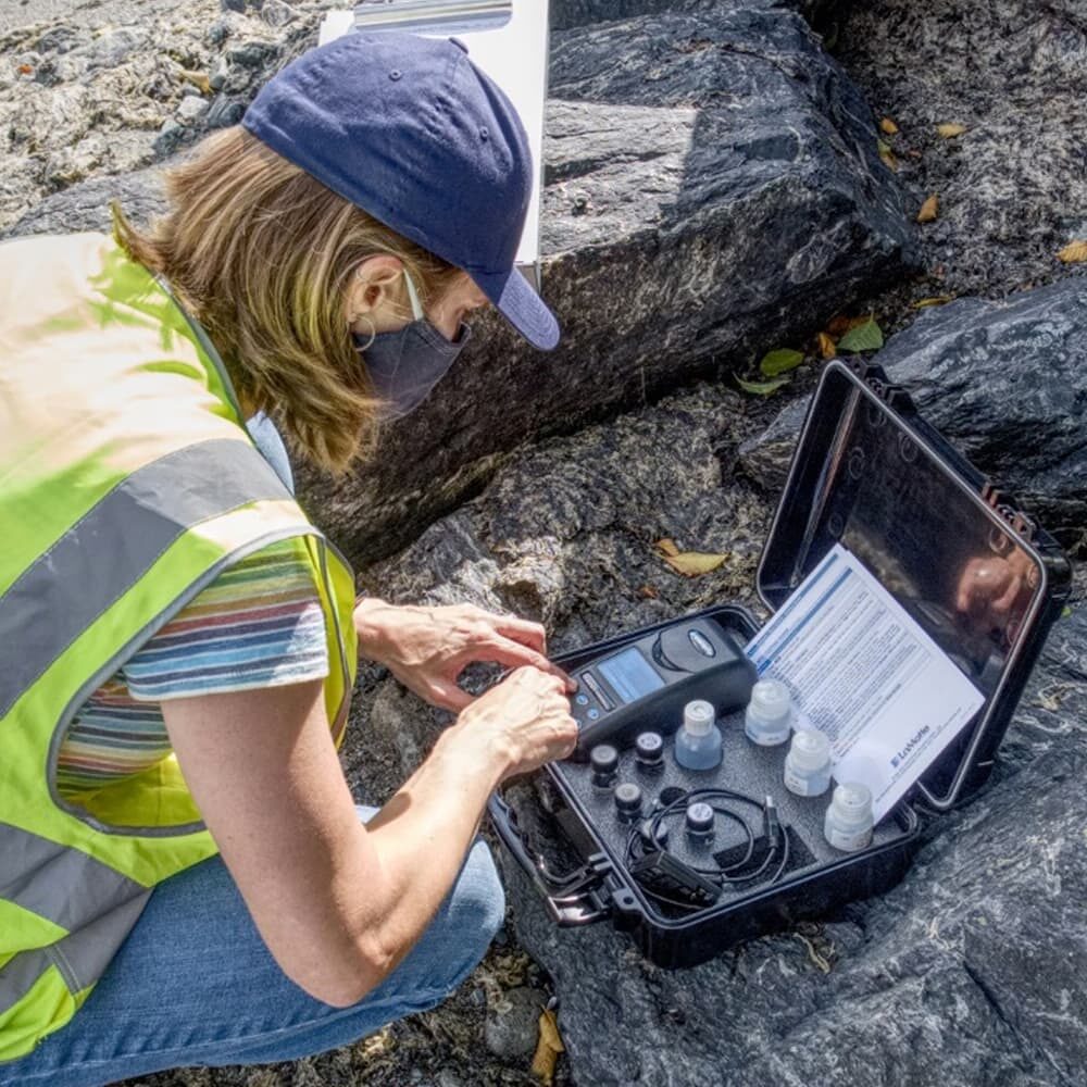 A person in a high-visibility vest and a navy cap is using scientific equipment in a black case on rocky terrain. They appear to be conducting field research or testing, with various instruments and bottles visible. They are also wearing a mask.