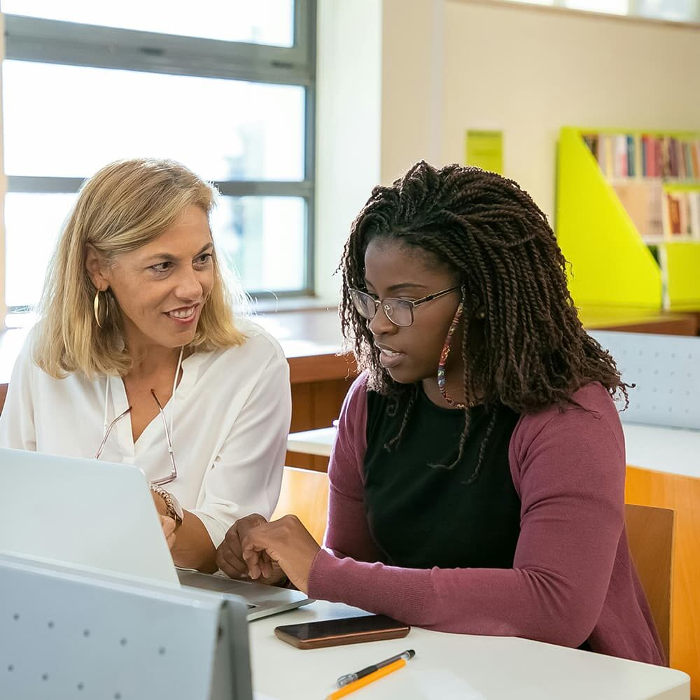 A woman with blonde hair and a white shirt sits beside a woman with long braided hair and glasses, wearing a black shirt and purple cardigan. They are looking at a laptop screen together in a brightly lit room with bookshelves in the background.