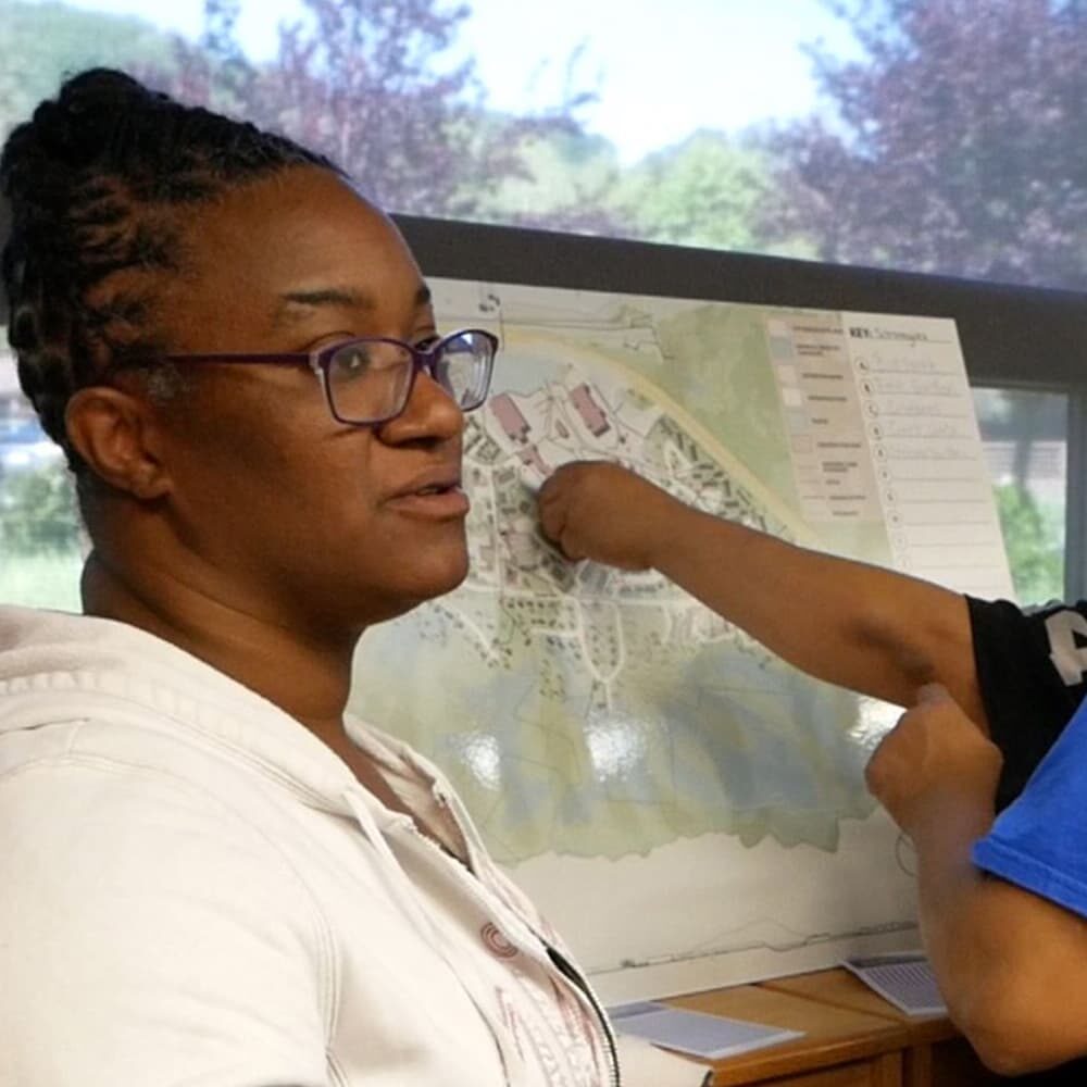 Una persona con gafas y sudadera blanca con capucha está de pie delante de un gran mapa. El brazo de otra persona, con una manga azul y negra, señala una zona concreta del mapa. Ambos individuos parecen mantener una discusión.