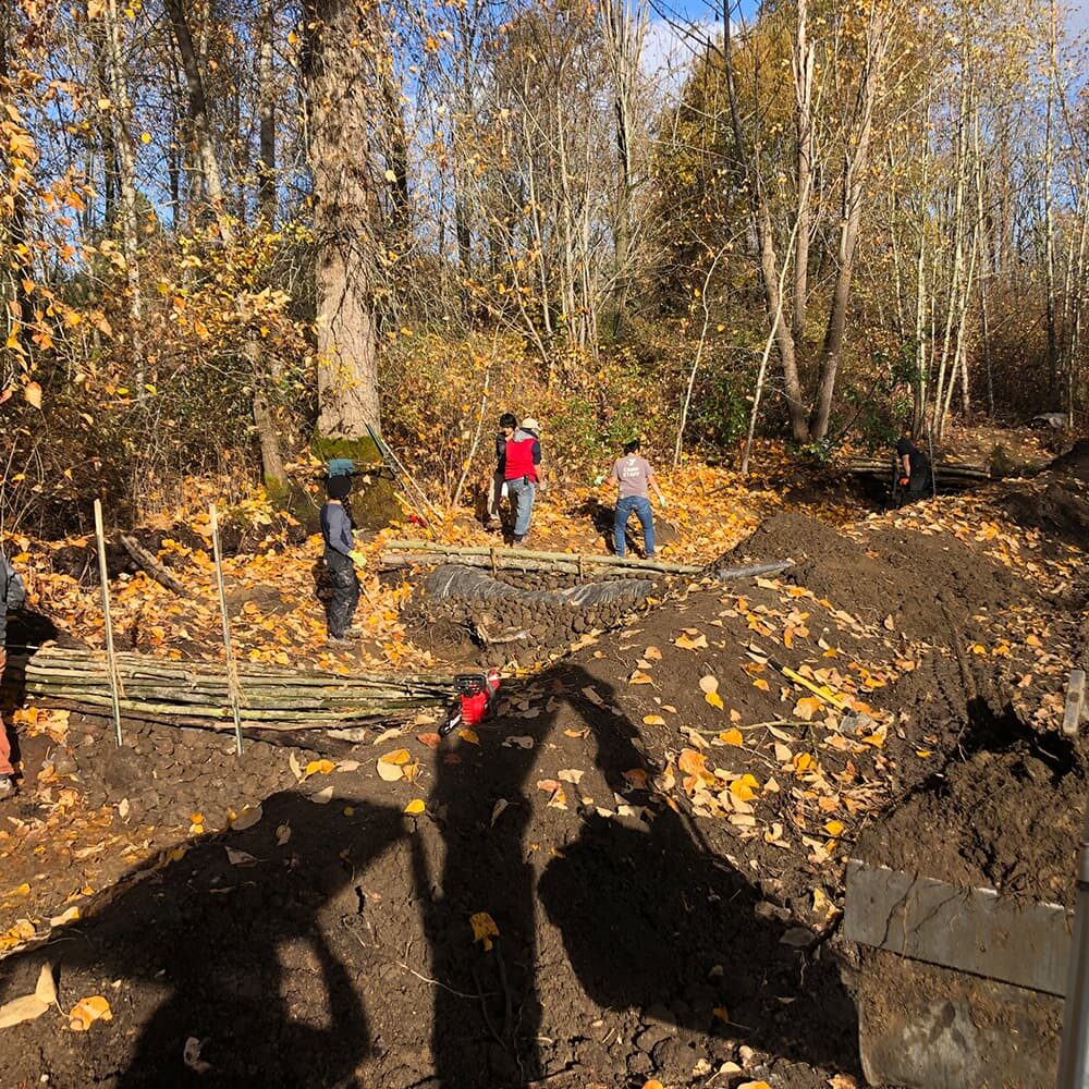 Several people work on a construction site in a forested area during autumn. They are surrounded by trees with colorful fall leaves. Coils of wire and a partially constructed structure are visible on the ground. A shadow of a person taking the photo is in the foreground.