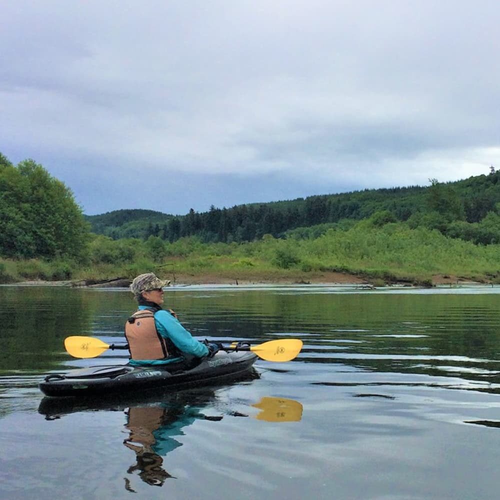 A person wearing a life jacket and cap is sitting in a kayak on a calm lake, holding a double-bladed paddle. The landscape features lush green trees and hills under a cloudy sky, reflecting beautifully on the water's surface.