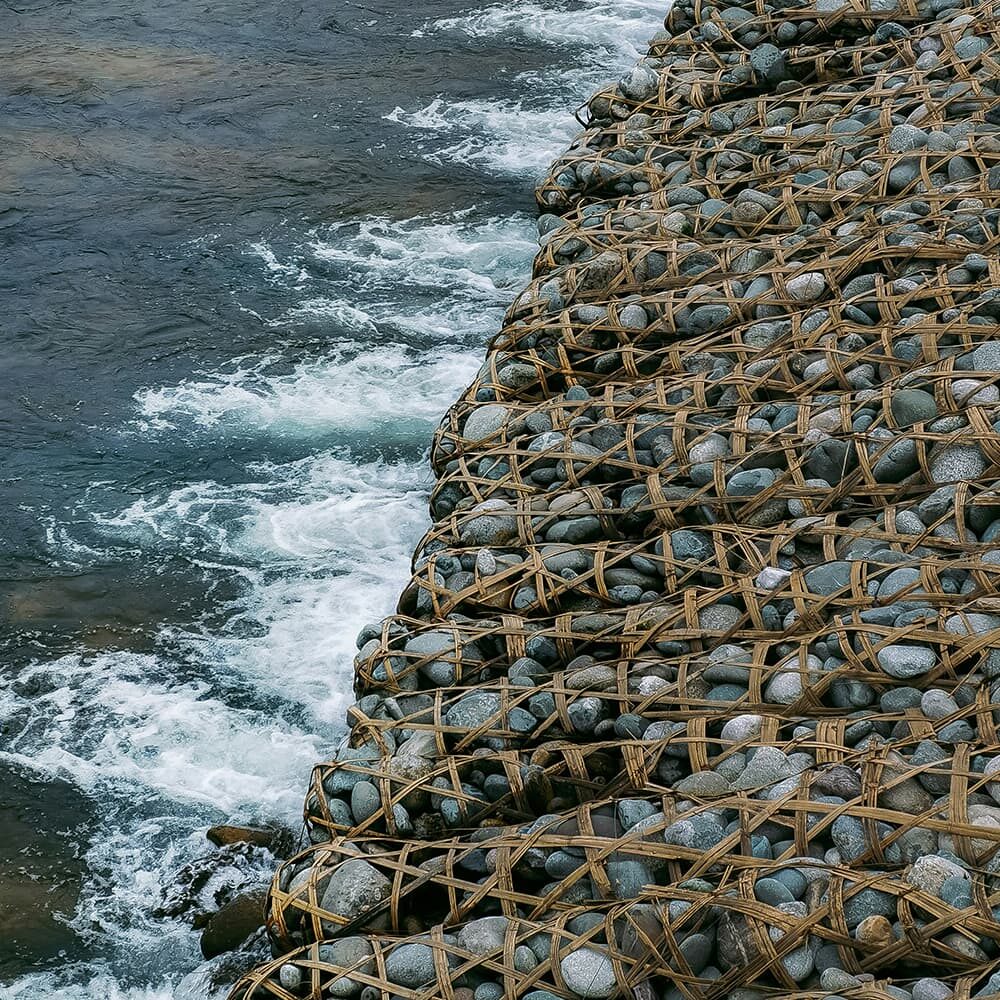 A section of a shoreline where smooth, rounded stones are contained by a woven rope net. The stones form a structured barrier next to the water, which is frothy with small waves splashing against the rocky edge. The image captures the texture and interaction of natural elements.