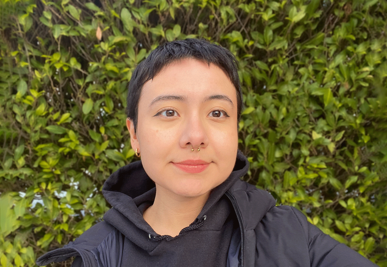 Headshot of a person with short black hair in a black jacket in front of a background of green leaves