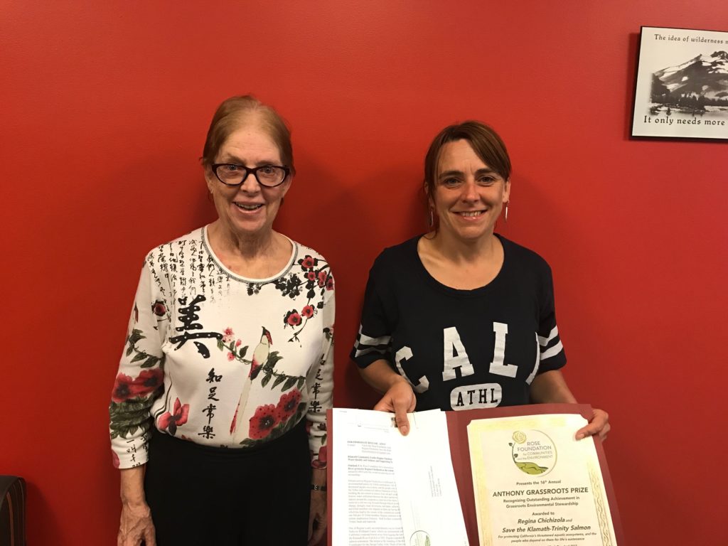 Two people standing in front of a red wall, one holding an open folder displaying a certificate.