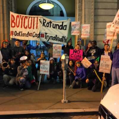 A group of people holding signs that read "Boycott The Rotunda" and "No Coal" are gathered in front of a building. The group appears to be participating in a protest. A large banner behind them features some of the same slogans. It is nighttime.