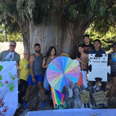 A group of people stands under a large tree, holding various signs related to climate change. The signs include colorful handprints, a wheel with text, and banners that read "Climate is a youth issue" and "No coal companies wanted". They appear to be at a protest or rally.