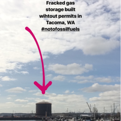 A large gas storage tank is labeled with text, "Fracked gas storage built without permits in Tacoma, WA #notofossilfuels," with a pink arrow pointing to it. The tank is near a marina with boats docked under a partly cloudy sky.