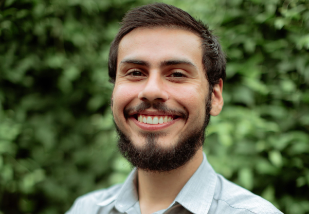 Headshot of a man with brown hair and beard in a collard shirt standing in front of a wall of green leaves
